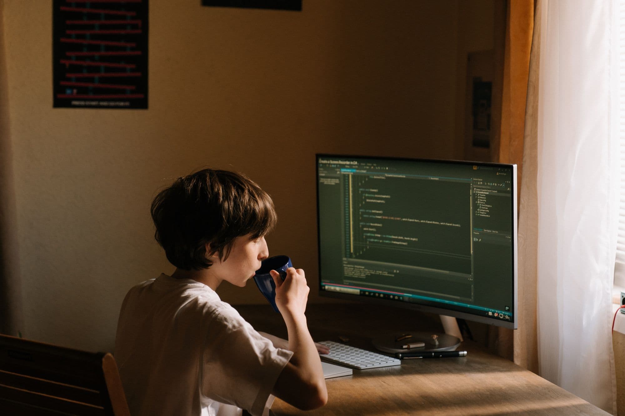 Person sitting in a sunlit room, sipping coffee in front of a curved computer monitor showing code