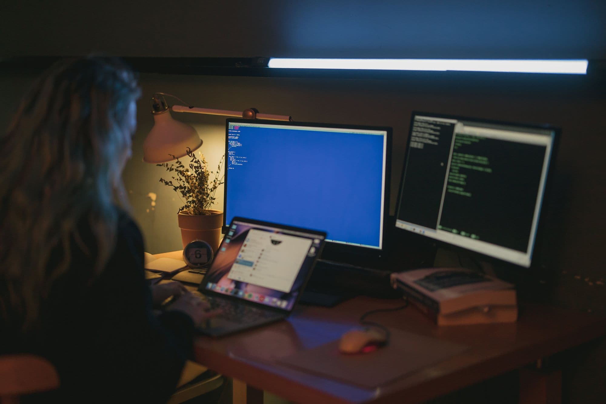 Woman sitting in a dark room in front of three monitors showing code